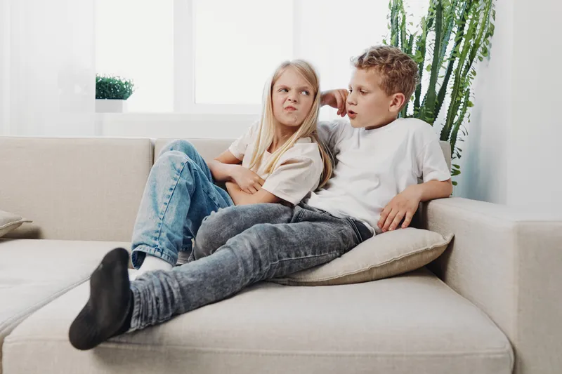 Two frustrated children sit on beige sofa indoors, girl crosses arms looking upset, boy talks with serious expression. Concept for sibling conflict, family tension, childhood emotions.