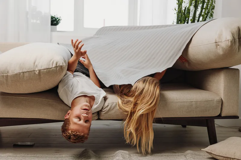 Two children play hiding under a blanket on beige sofa in bright living room, enjoying playful moment and family fun indoors together.