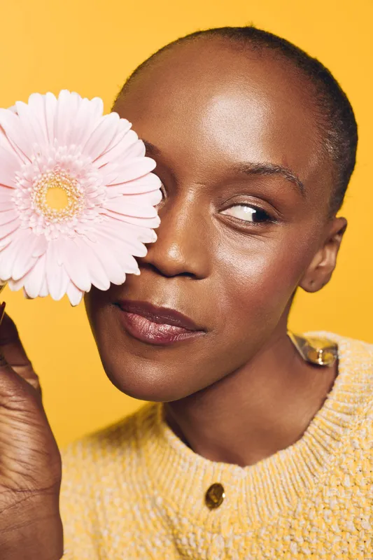 Close-up of a dark skinned African woman holding a pink flower near her face against a yellow background. Concept of natural beauty, skincare, and floral wellness with bright studio lighting.