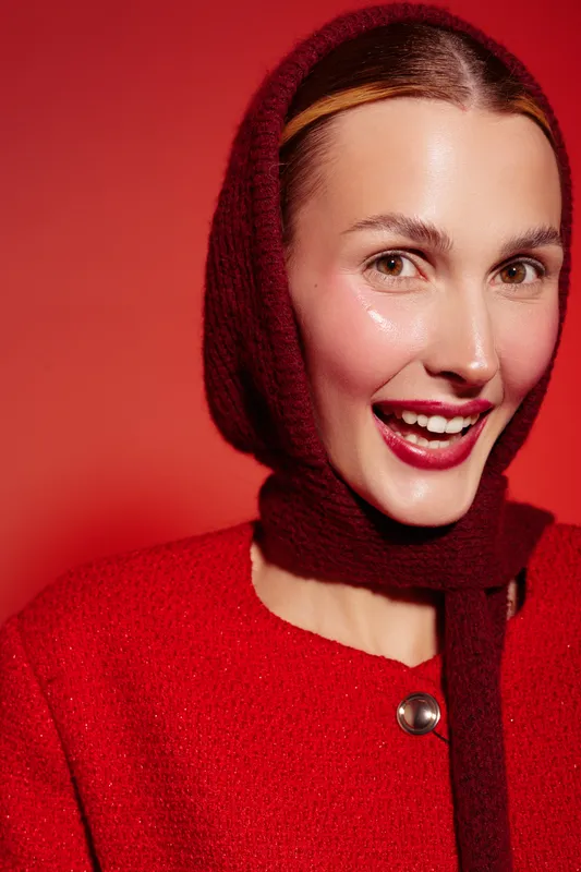Smiling woman in red woolen scarf and coat poses against red background. Close-up portrait with bright makeup and glossy skin. Concept of fashion, winter style, and beauty.