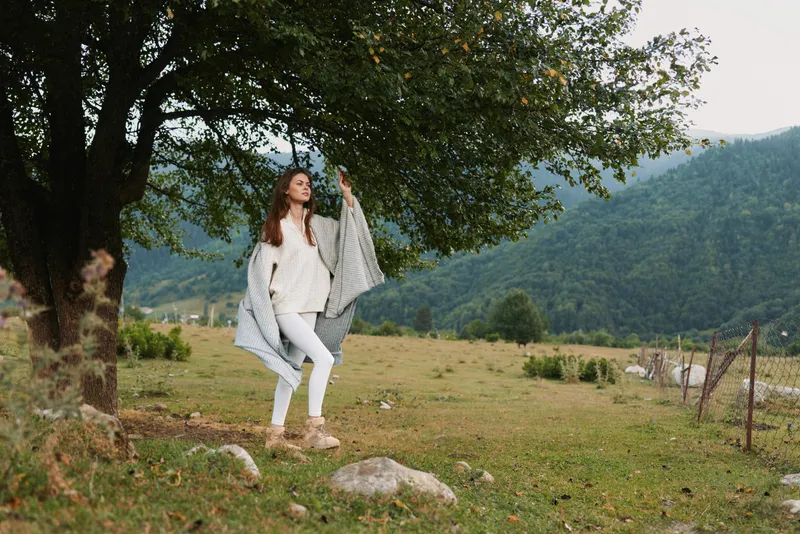 A young woman stands beside a large tree in a quiet meadow, wearing a white outfit and a light shawl, gazing upward with a calm, hopeful expression under a shaded, open sky.