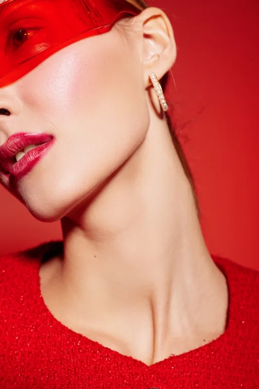 Close-up of a woman wearing a red translucent mask and red sweater, showing smooth skin and gold hoop earrings. Concept of fashion, beauty, and modern style on red background.