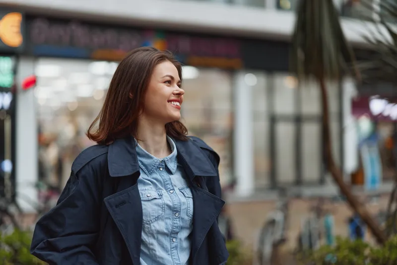 Cheerful woman outdoors, smiling in a denim jacket, city street backdrop, natural light, casual pose, light breeze, vibrant colors, urban atmosphere