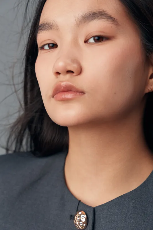 Portrait of young Asian woman with natural makeup and long black hair wearing grey garment with decorative button standing against plain background with confident expression.