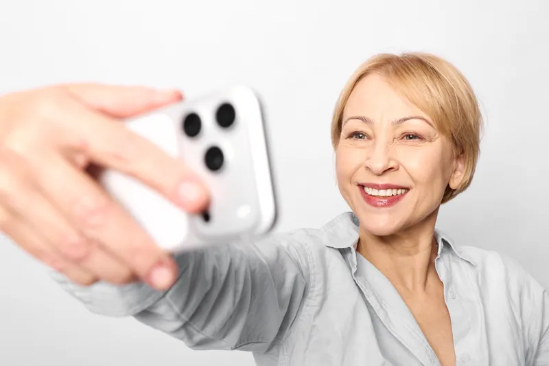 Woman selfie smartphone senior smiling technology communication social media, elderly woman holds white smartphone for selfie, bright studio background, cheerful.