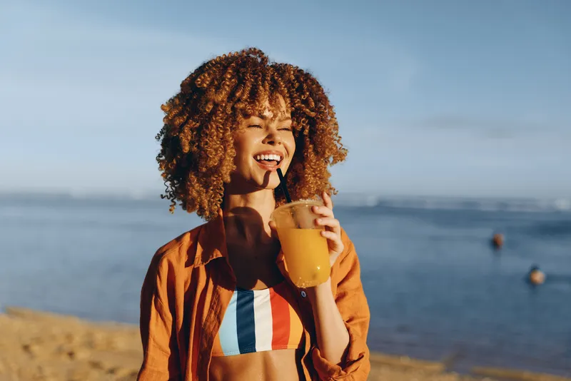 Woman with curly hair smiles near ocean, holding yellow mug of warm drink, casual striped jacket, golden outdoor sunshine, calm seaside lifestyle, blurred horizon
