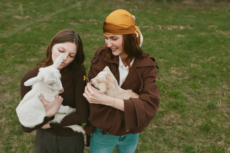 A pair of women share a gentle moment with a baby goat in an open grassy field, capturing a wholesome lifestyle scene focused on animal care, friendship, and outdoor farm life.