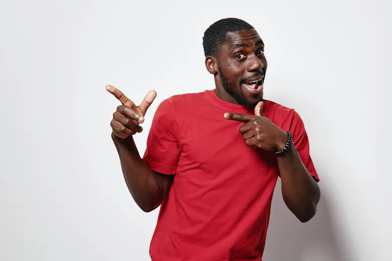 African American man with dark skin wearing red t shirt smiling and pointing fingers sideways on white background. Happy male portrait with casual clothing and positive expression.