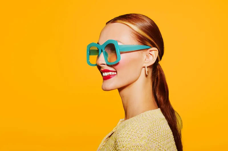 Young woman with red hair and bright red lipstick wears large turquoise sunglasses and yellow sweater against vibrant yellow background. Concept of fashion, style, and summer mood.