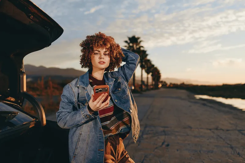 Woman lifestyle, curly hair, denim jacket, holding smartphone by open car, walking on road at sunset, outdoor portrait with warm evening light, relaxed, smiling softly, streetwear, casual