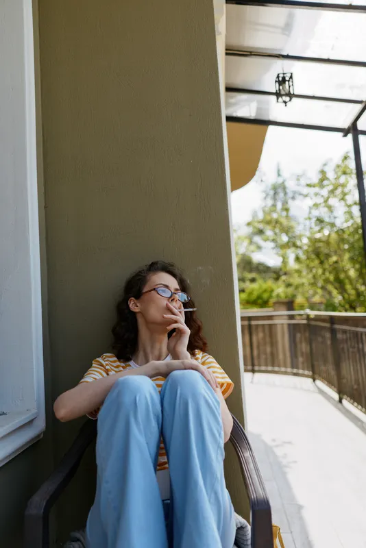 Relaxed woman on a balcony smoking a cigarette, seated in a casual striped shirt and blue pants. Outdoor light creates a calm mood, home lifestyle and quiet contemplation.