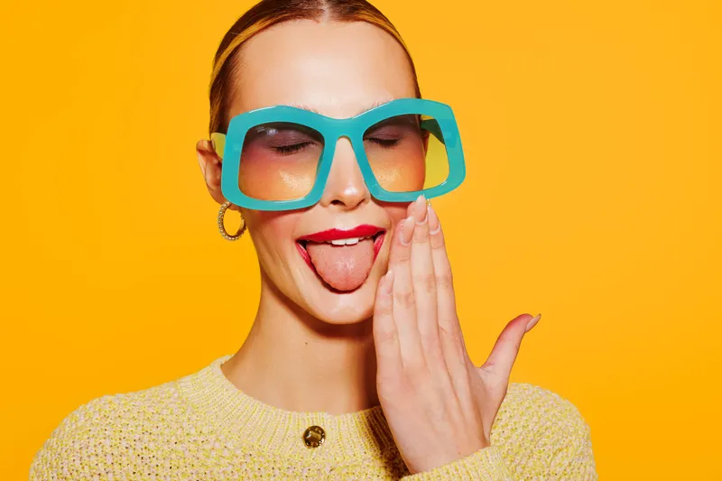 Young woman with large blue glasses and yellow sweater makes funny face sticking out tongue on bright orange background. Concept of playful mood, fashion, and colorful portrait.