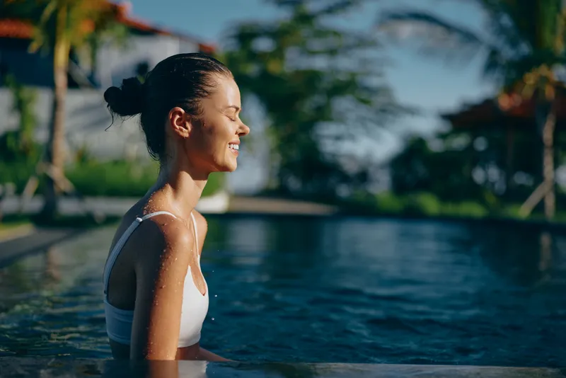A young woman in a white swimsuit relaxes in a pool with eyes closed, enjoying the sunny day and peaceful outdoor environment. Concept of summer lifestyle and wellness.