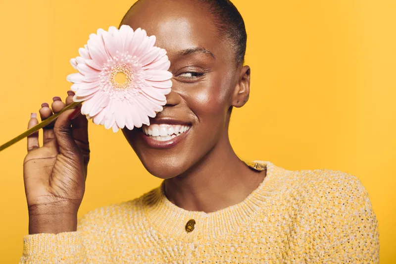 Happy dark skinned African woman smiles holding pink flower near face on yellow background. Concept of natural beauty, joy, skincare, wellness, and positive emotions in studio lighting.