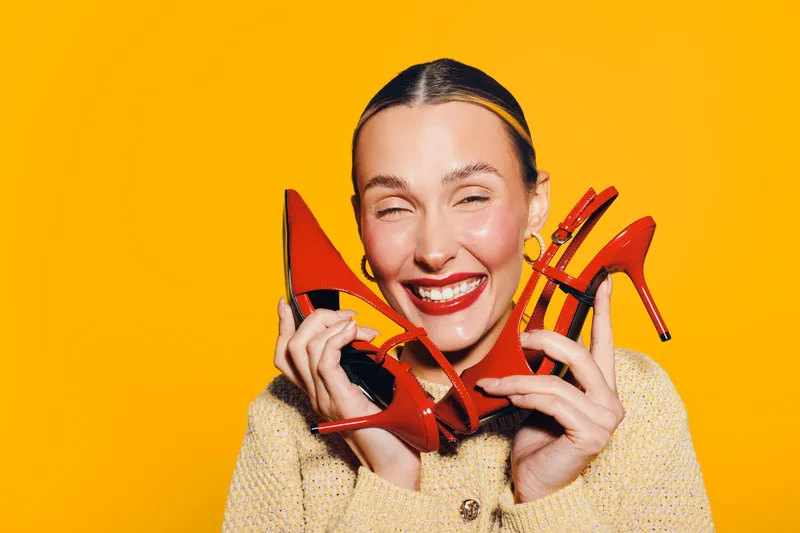 Happy woman with bright red lipstick holds red high heel shoes near face smiling with closed eyes on yellow background. Concept of fashion, style, and joyful emotions.