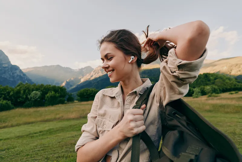 Happy woman outdoors hiking in mountains, smiling with earbuds, casual outfit, enjoying nature, green fields and blue sky, active lifestyle moment