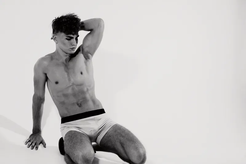 Young man with muscular body posing in underwear sitting on floor with hand behind head in studio with white background and soft lighting for fitness concept.