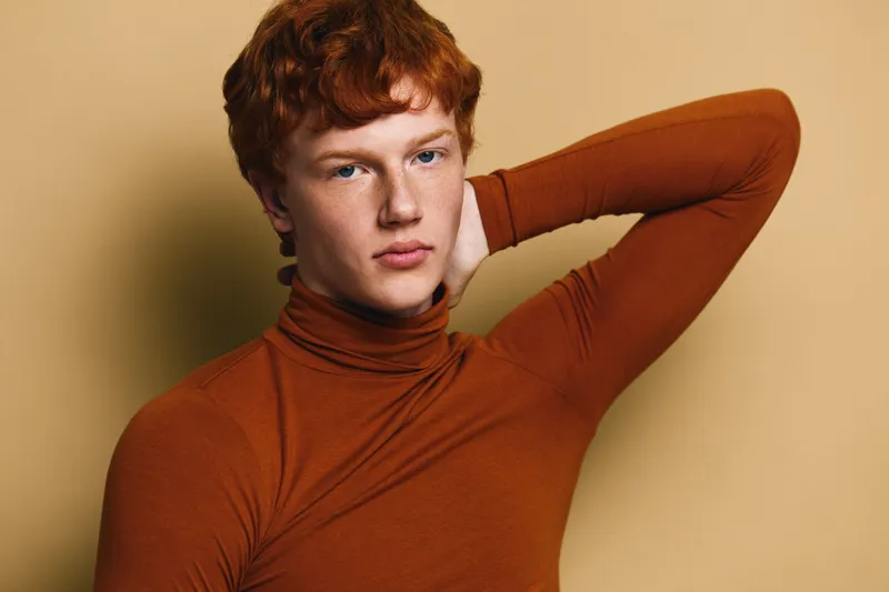 Young man with red hair and blue eyes posing with hand behind neck wearing brown turtleneck sweater against beige background. Closeup portrait of thoughtful male model in studio.