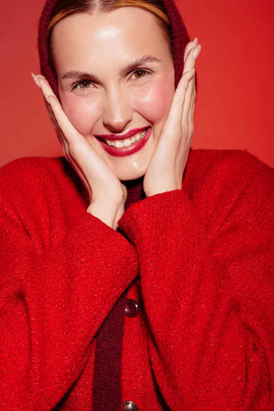 Smiling woman in red sweater poses with hands on face against red background. Close-up portrait with natural makeup and glossy skin. Concept of happiness, fashion, and beauty.