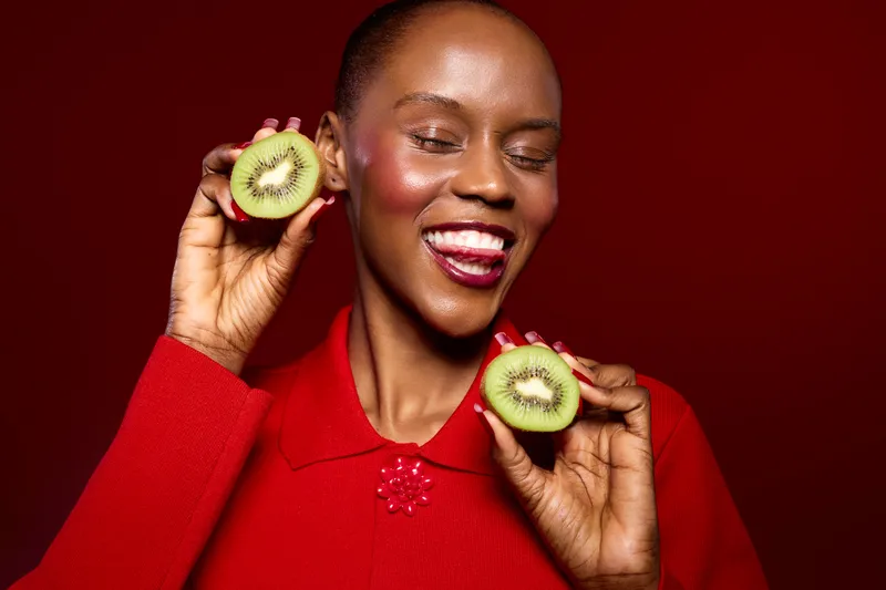 Happy African woman with dark skin wearing red sweater holds two halves of kiwi fruit near face with eyes closed and smiling. Concept of healthy food, natural beauty, and joyful lifestyle.
