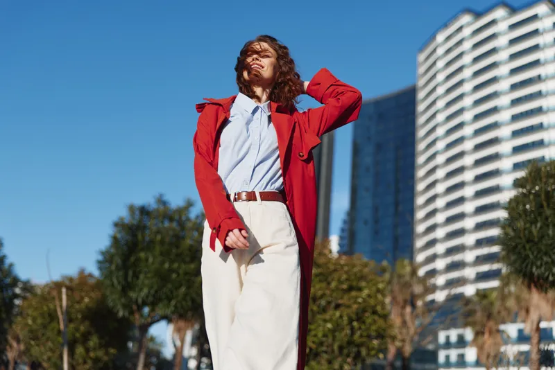 Young adult with wavy brunette hair wears a bright red coat over a white shirt and beige trousers, standing confidently in an urban plaza under a clear blue sky.