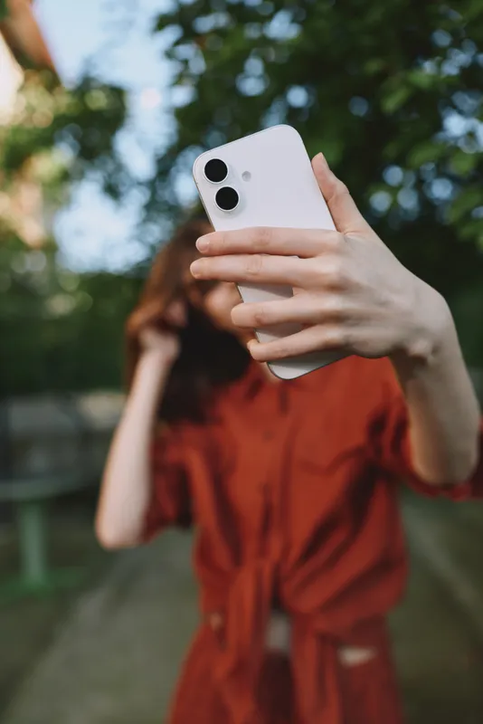 Young woman in a red dress outdoors takes a selfie with a white smartphone, smiling softly as lush greenery frames her in a sunlit park