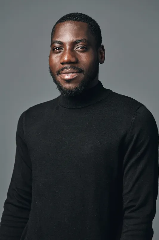African American man with dark skin wearing black turtleneck sweater posing with confident smile in creative visual designer style studio portrait on gray background.