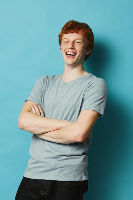 Young man with red hair smiling in grey t shirt standing with crossed arms against blue background for casual portrait and happiness expression in studio.