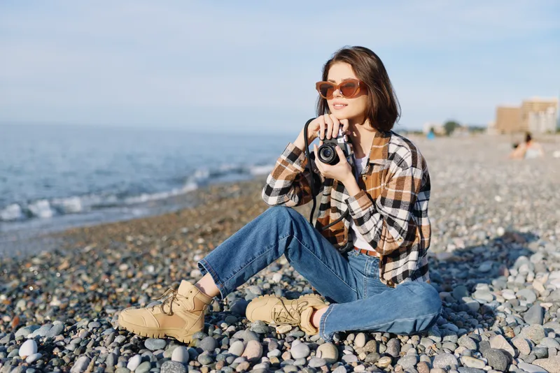beach photographer camera outdoor casual lifestyle sea pebbles sunglasses coastal calm shore moment sunlight capturing landscape with a woman on a pebble beach wearing plaid shirt jeans boots and a