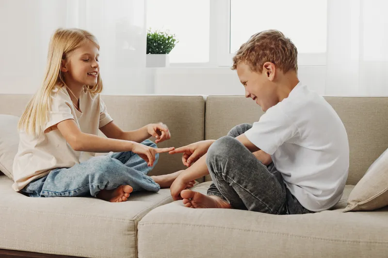 Happy young boy and girl sitting on beige sofa playing finger game in bright living room with natural light and casual clothes.