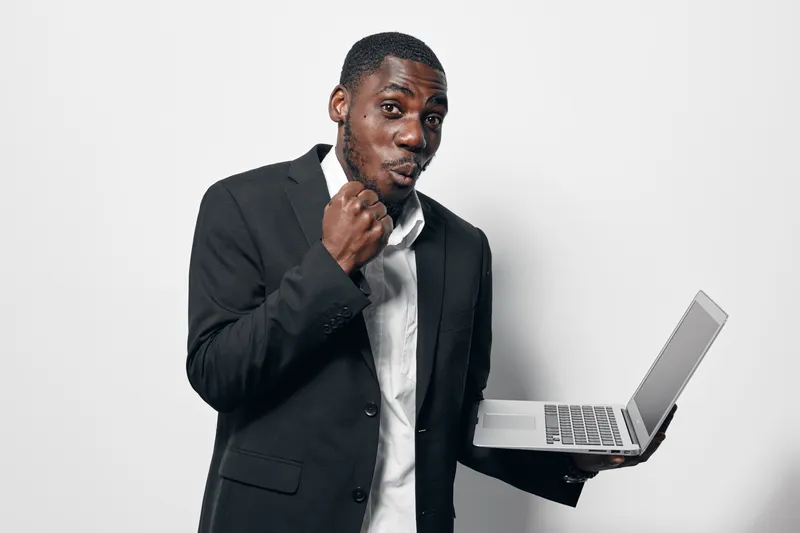 African American man with dark skin in black suit holding laptop and making fist gesture for success or excitement on white background. Business professional with confident expression.