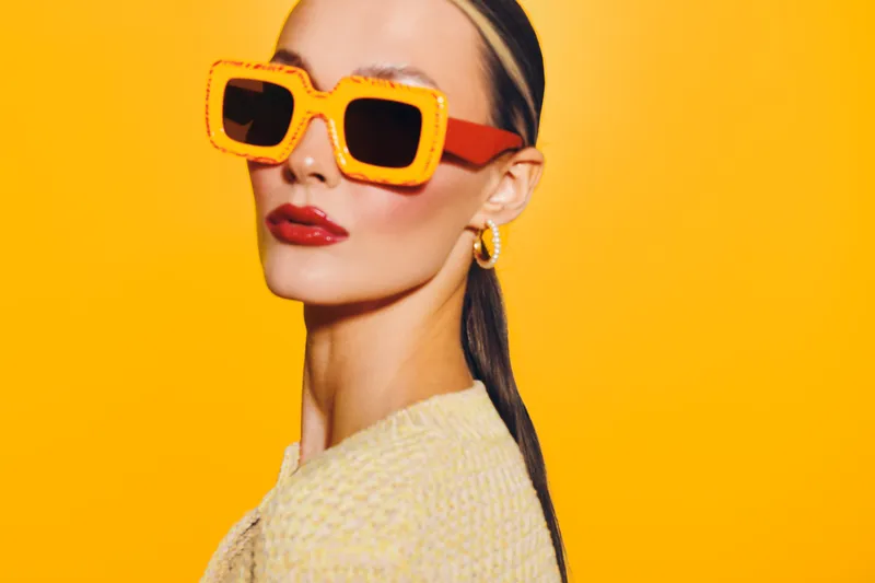 Stylish woman with slicked back hair wears oversized orange sunglasses and red lipstick against bright yellow background. Fashion portrait with bold colors and confident expression.