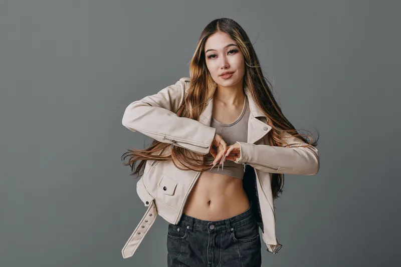Young woman with long brown hair wearing beige jacket and gray crop top posing with hand gesture on gray background. Casual style, confident and modern female portrait.