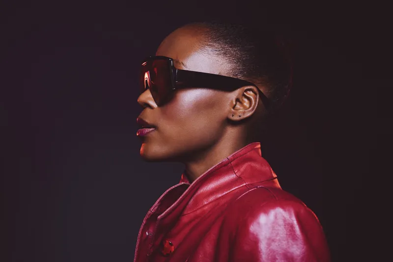 Profile of a confident African woman with dark skin wearing large sunglasses and a red leather jacket against a dark background. Concept of fashion, style, and modern beauty.