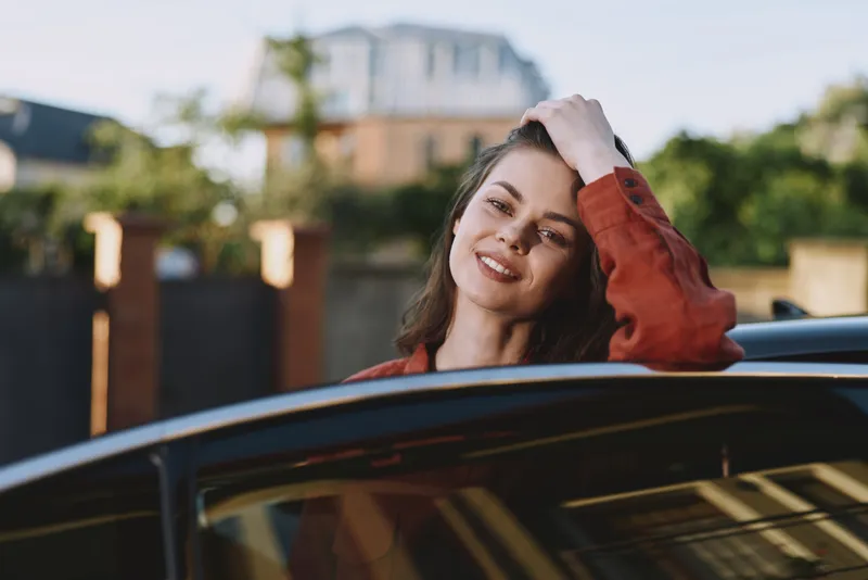 Woman smiles from car window during daytime, casual lifestyle portrait, relaxed mood, outdoor suburban scene, warm natural light, candid moment of freedom and joy, captured authentically