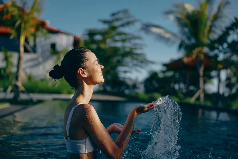 Young woman in a white swimsuit creates a spray of water in a sunlit pool, highlighting outdoor leisure, summer vitality, tropical atmosphere, sunshine, and a carefree mood that conveys health,
