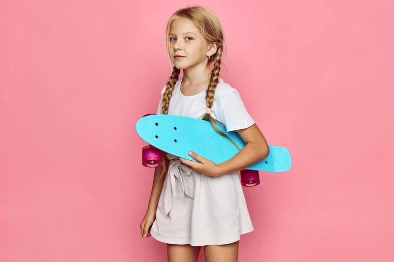 Blonde girl with two braids holds a blue skateboard while standing against a pink background. She wears a white shirt and beige skirt with a confident expression.