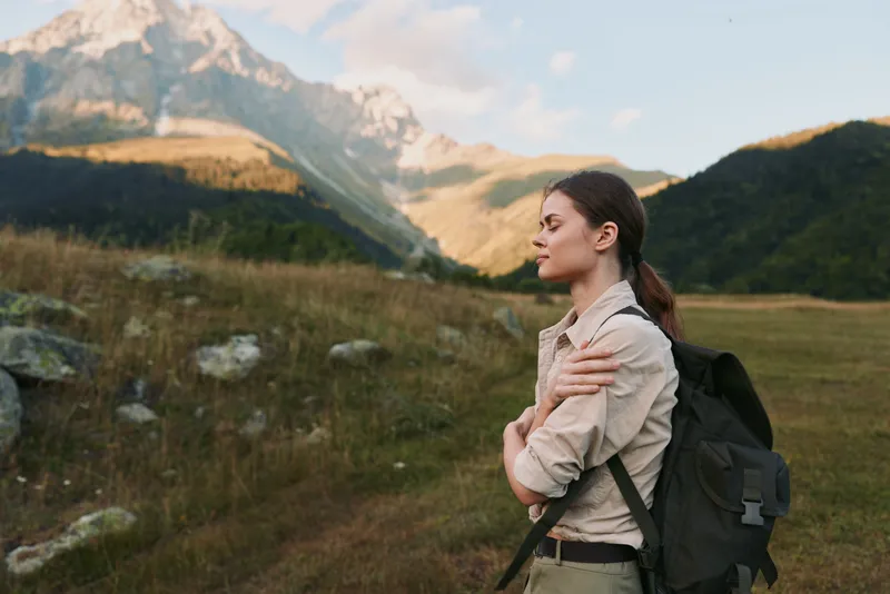 Young woman with a backpack gazes toward distant mountains in a sunlit valley, embodying exploration, calm focus, and a sense of freedom in the open landscape.
