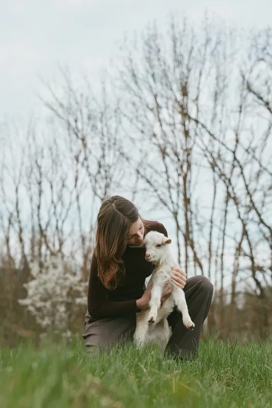 Young woman in a black sweater lovingly holds a white baby goat while sitting on the grass in a natural outdoor setting during early spring.