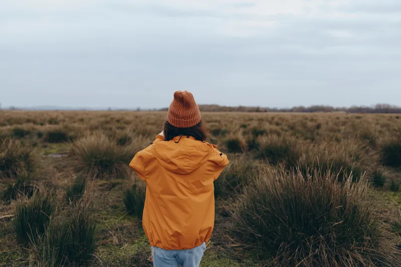 Woman outdoors, lifestyle in a field of grass, hooded jacket and knit hat, walking away along a dirt path under cloudy sky, calm rear view, soft natural tones