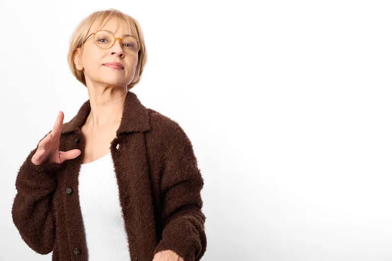 elderly woman with glasses, brown coat portrait on white background, raises hand in friendly gesture, calm smile, concept for senior communication and lifestyle studio
