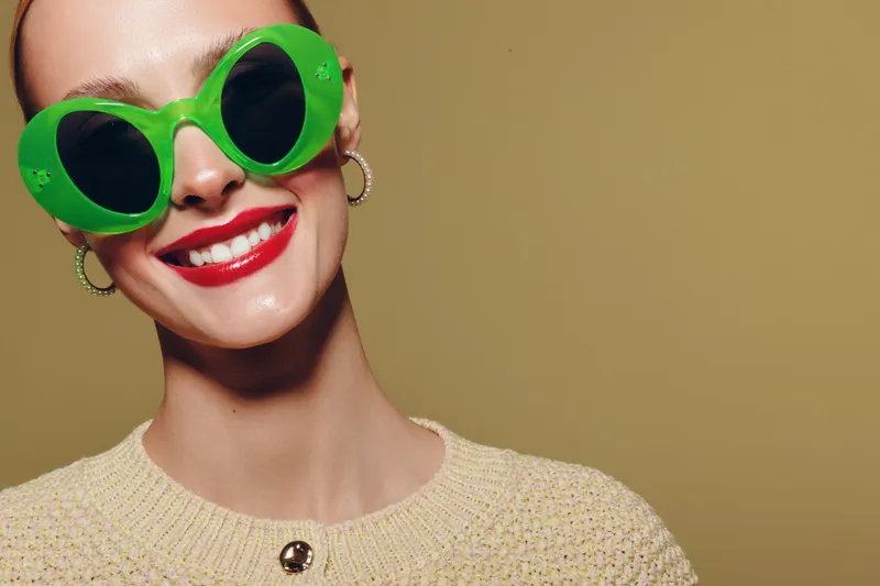 Smiling woman with bright red lipstick wears large green sunglasses and beige sweater. Close-up portrait on yellow background. Concept of fashion, style, and summer accessories.