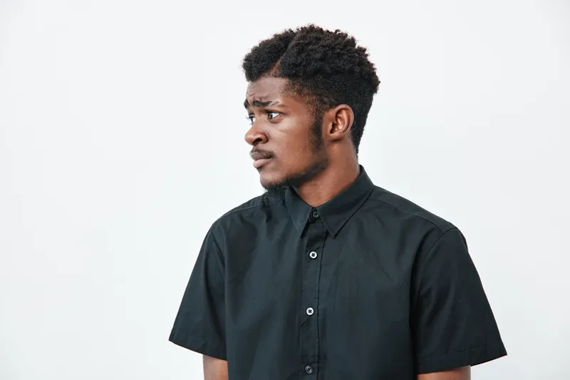 Young black man with dark skin wearing black shirt looking sideways on white background. Portrait of thoughtful African American male with short curly hair and casual style.