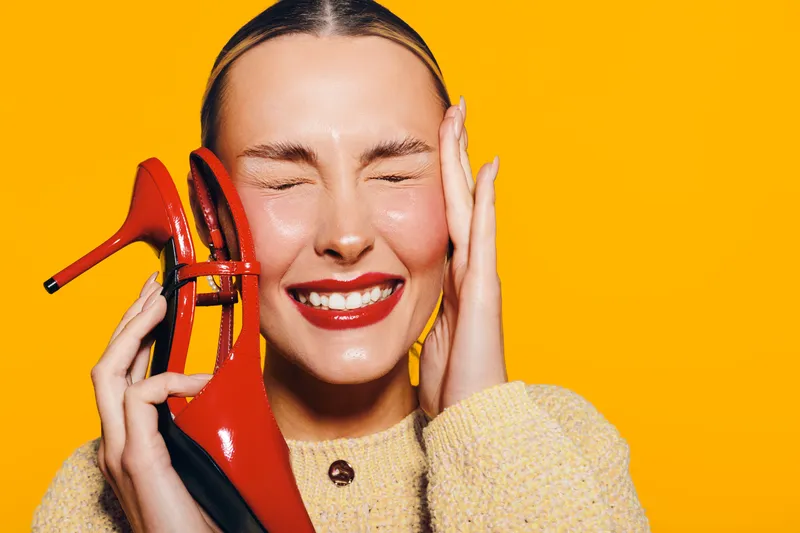Happy woman with red lipstick holds red high heel shoe near face, eyes closed, smiling. Bright yellow background and cozy sweater create warm, joyful mood. Concept of fashion and happiness.