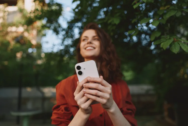 A cheerful woman outdoors holds a smartphone, looking up with a warm smile. Bright daylight and lush greenery frame a casual, confident lifestyle moment