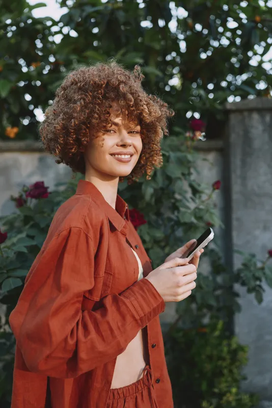 Cheerful woman outdoors, smiling while looking at her smartphone, casual rust outfit, natural light, leafy background, relaxed lifestyle, candid moment