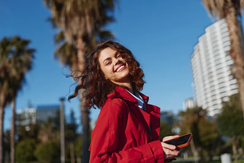 Young woman with curly hair smiles joyfully outdoors wearing a red jacket while holding a smartphone on a sunny day with palm trees in the background.