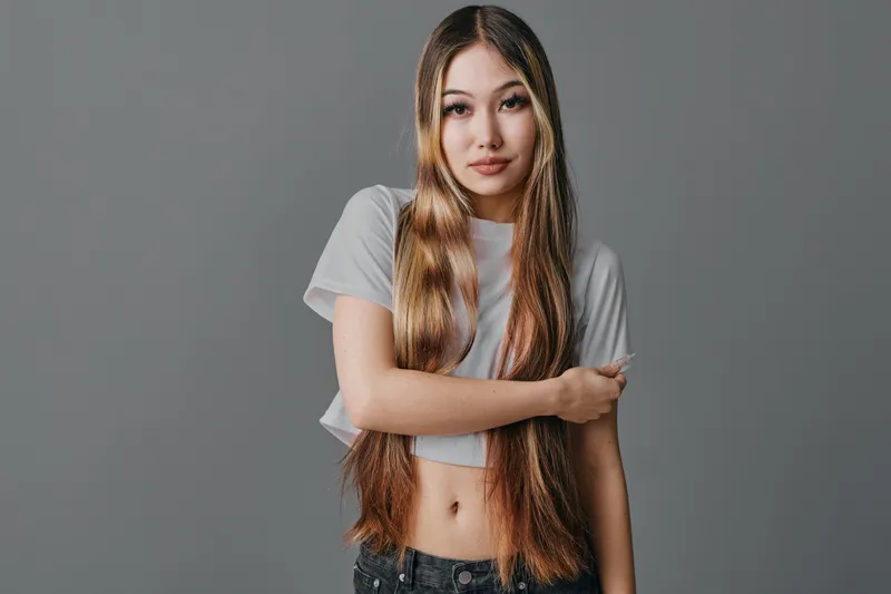 Young woman with long straight hair wearing casual white t-shirt and black jeans posing with arms crossed against gray background in studio portrait.