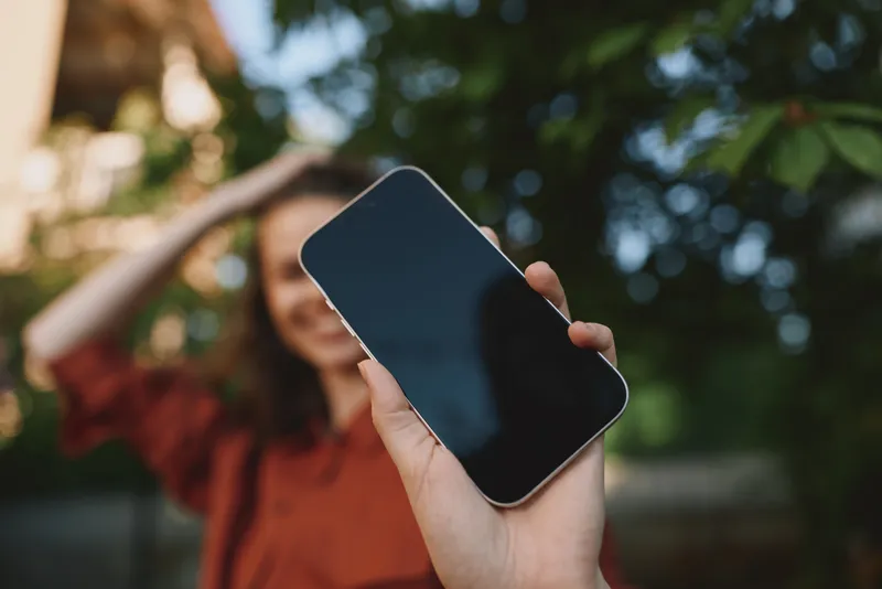 Smiling woman outdoors holds a smartphone toward the camera, capturing a casual lifestyle moment. Bright daylight, green background, relaxed mood, and natural candid photography
