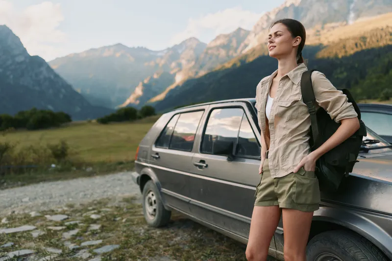 Woman traveler rests beside a car in a rugged mountain landscape, wearing casual hiking clothes and a backpack, enjoying fresh air and expansive scenery.